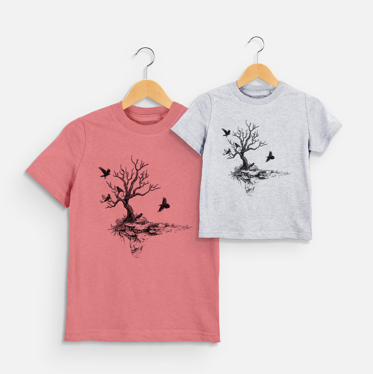 Two youth t-shirts in different colors on hangers, printed with a black ink illustration of a bare twisted tree on a small island with ravens flying around it.
