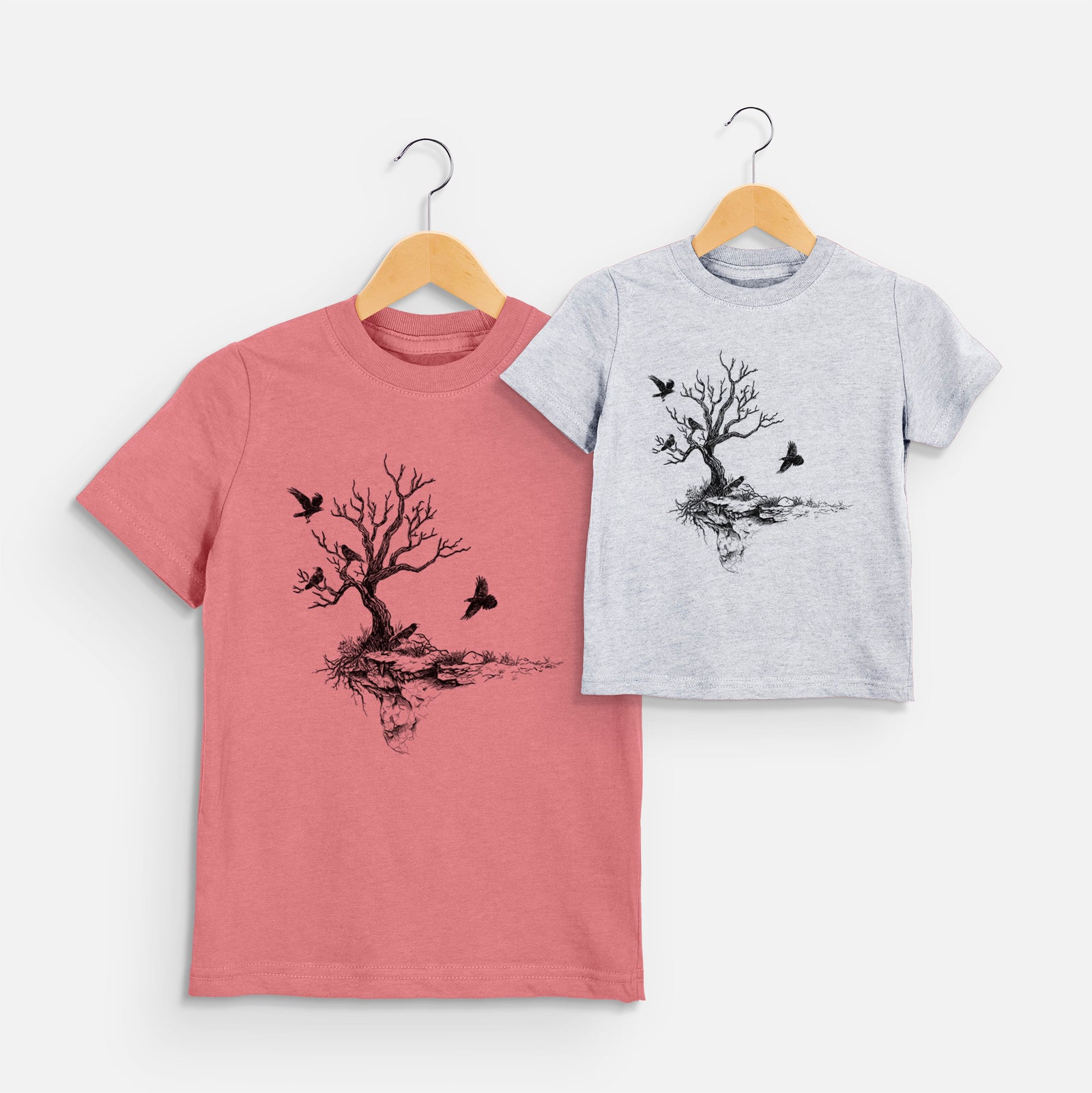 Two youth t-shirts in different colors on hangers, printed with a black ink illustration of a bare twisted tree on a small island with ravens flying around it.