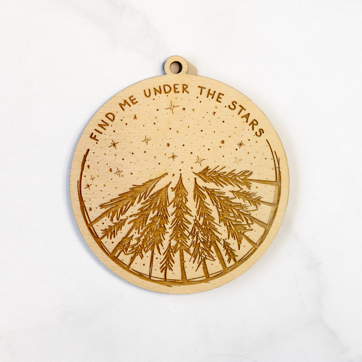 Wooden ornament featuring an engraved circle of pine trees under a starry sky, with the words Find Me Under the Stars, on a marble surface.