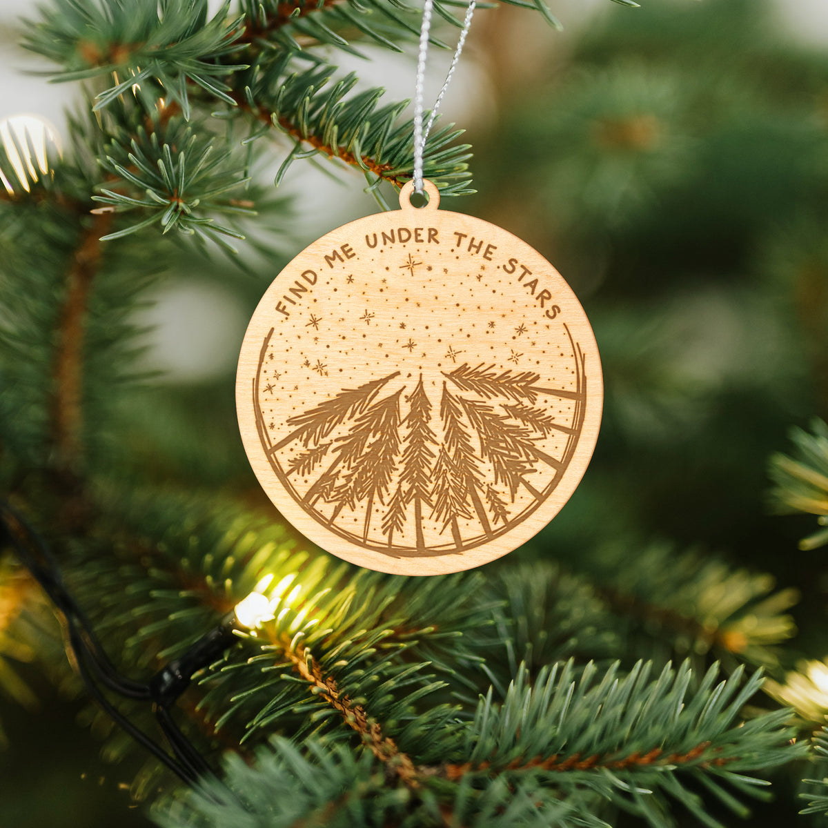 Wooden ornament featuring an engraved circle of pine trees under a starry sky, with the words Find Me Under the Stars, hanging on a Christmas tree.