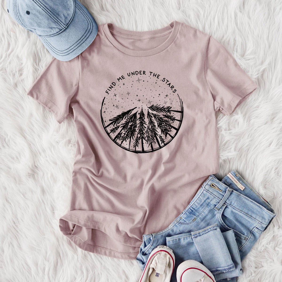 Women's relaxed fit cotton t-shirt printed with a circle of pine trees under a starry sky, with the words Find Me Under the Stars, styled flat on a white faux fur rug with jeans.