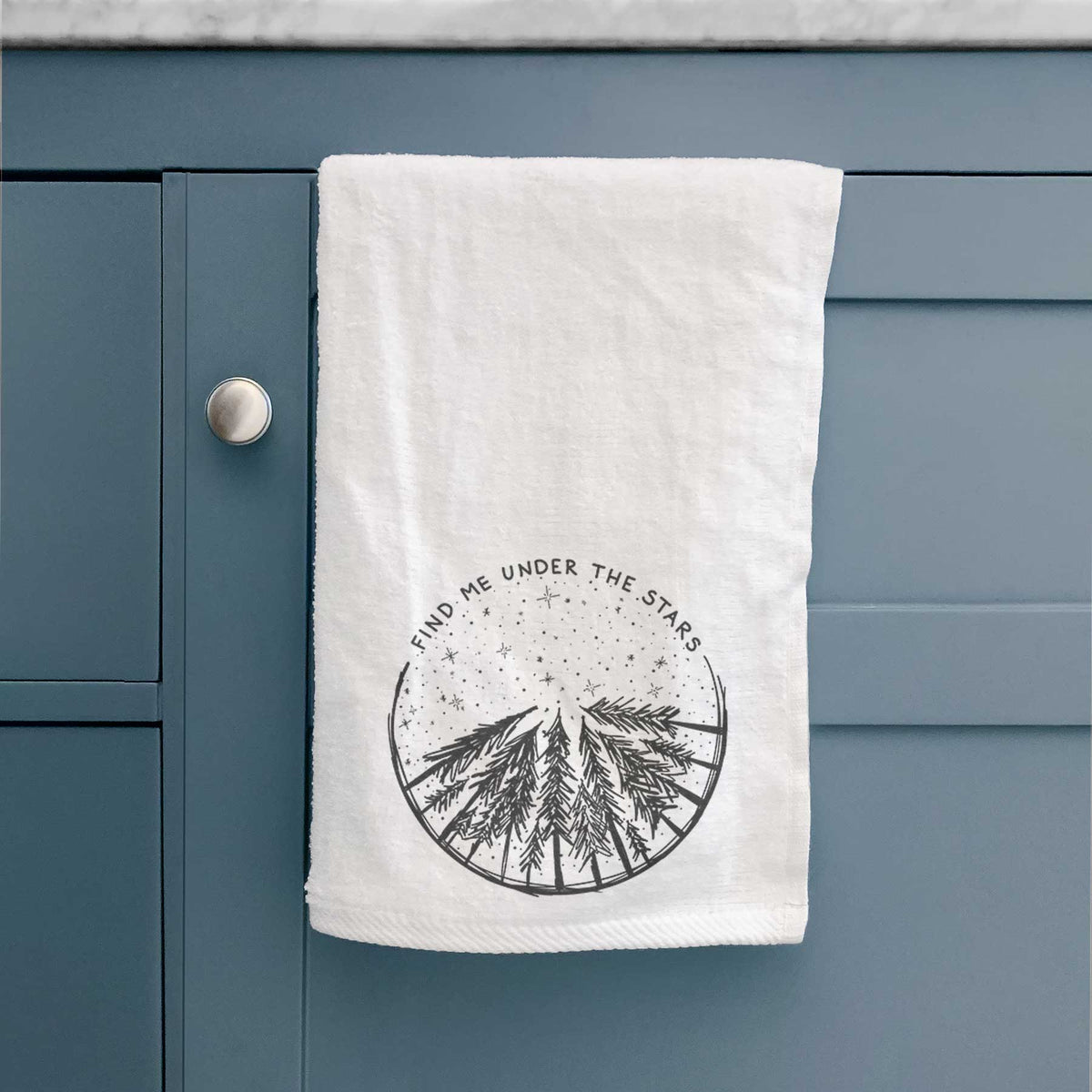 White hand towel printed with a circle of pine trees under a starry sky, with the words Find Me Under the Stars, hanging on a cabinet door.
