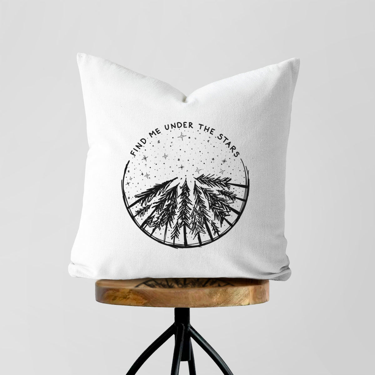 White pillow cover printed with a circle of pine trees under a starry sky, with the words Find Me Under the Stars, on a wooden stool.