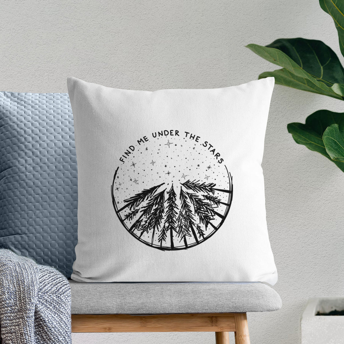 White pillow cover printed with a circle of pine trees under a starry sky, with the words Find Me Under the Stars, on a bench beside plants.