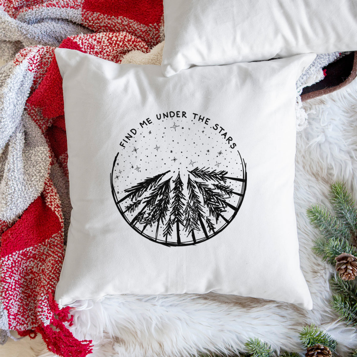 White pillow cover printed with a circle of pine trees under a starry sky, with the words Find Me Under the Stars, on a woven chair.