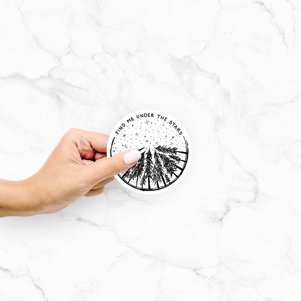 Hand holding a 4 inch vinyl decal of a circle of pine trees under a starry sky, with the words Find Me Under the Stars.