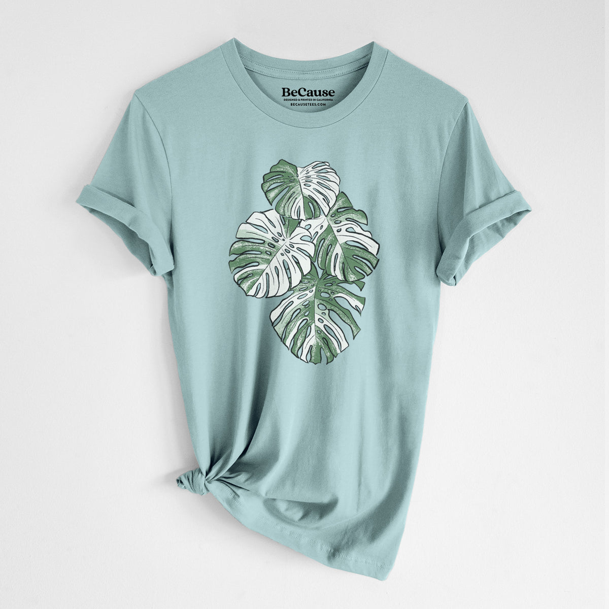 Dusty blue lightweight cotton t-shirt laid flat with the sleeves rolled, printed with a full-color illustration of a variegated Monstera leaf in green and white.