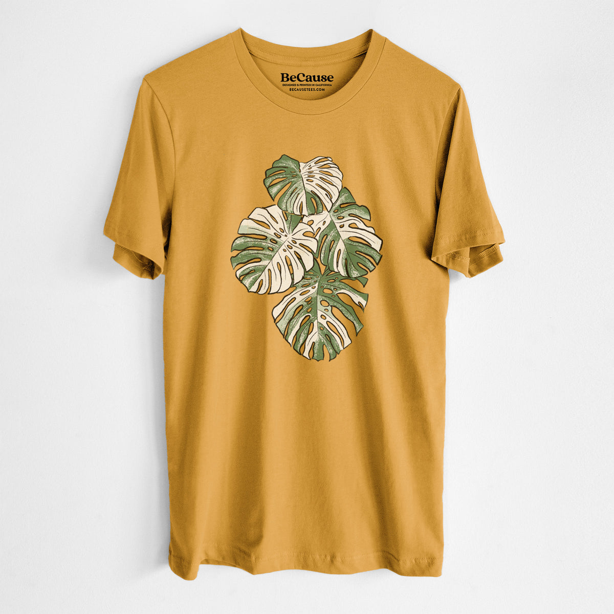 Mustard yellow lightweight cotton t-shirt laid flat, printed with a full-color illustration of a variegated Monstera leaf in green and white.