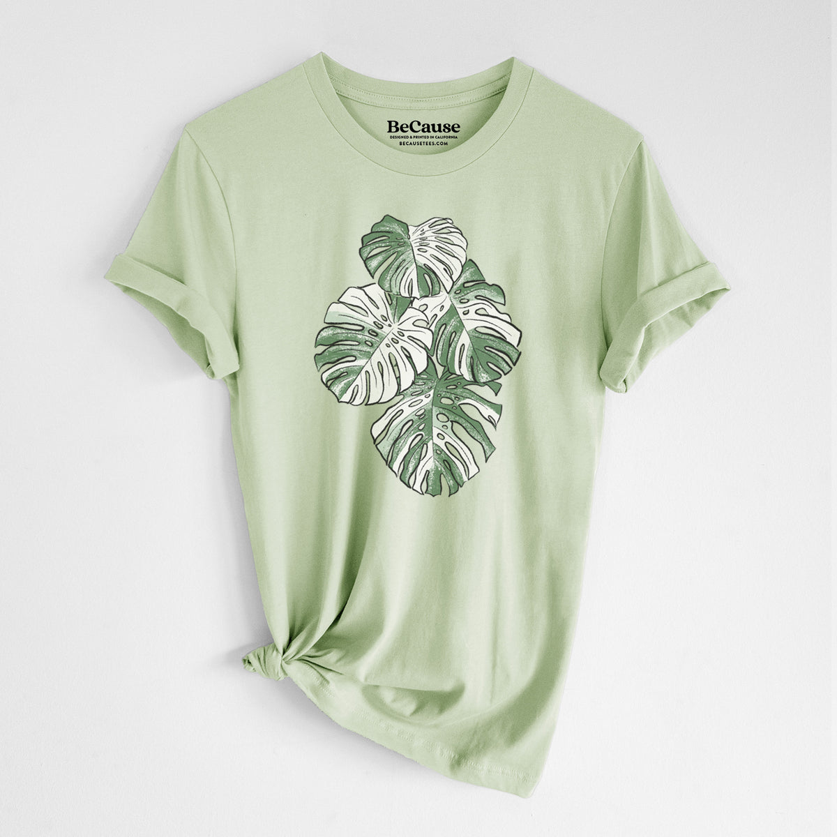 Spring green lightweight cotton t-shirt laid flat with the sleeves rolled, printed with a full-color illustration of a variegated Monstera leaf in green and white.