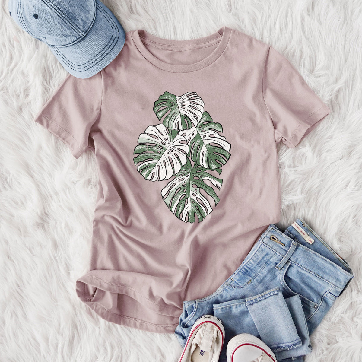 Women's relaxed fit cotton t-shirt printed with a full-color illustration of a variegated Monstera leaf in green and white, styled flat on a white faux fur rug with jeans.