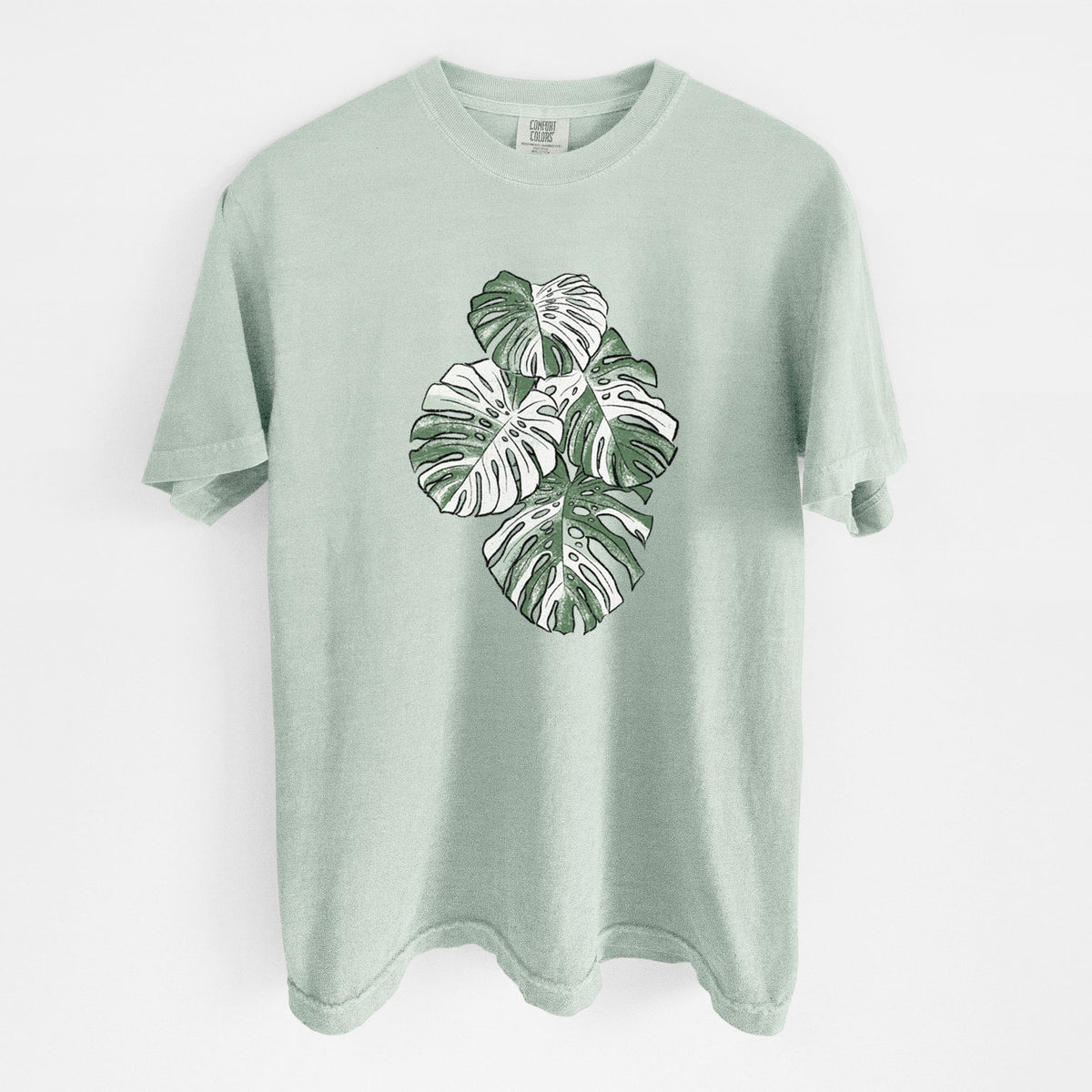 Bay green Comfort Colors t-shirt laid flat, printed with a full-color illustration of a variegated Monstera leaf in green and white.