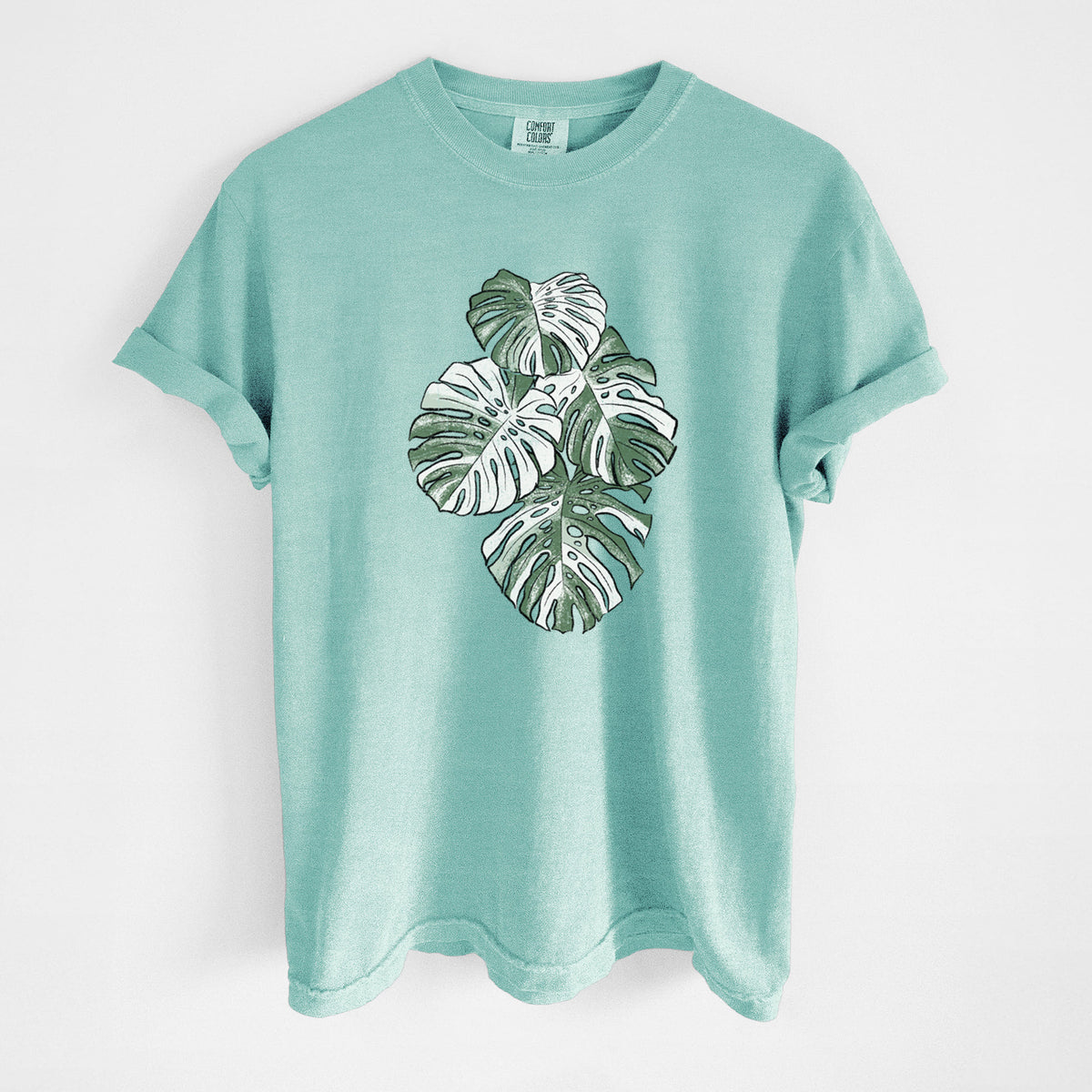 Chalky mint Comfort Colors t-shirt laid flat, printed with a full-color illustration of a variegated Monstera leaf in green and white.