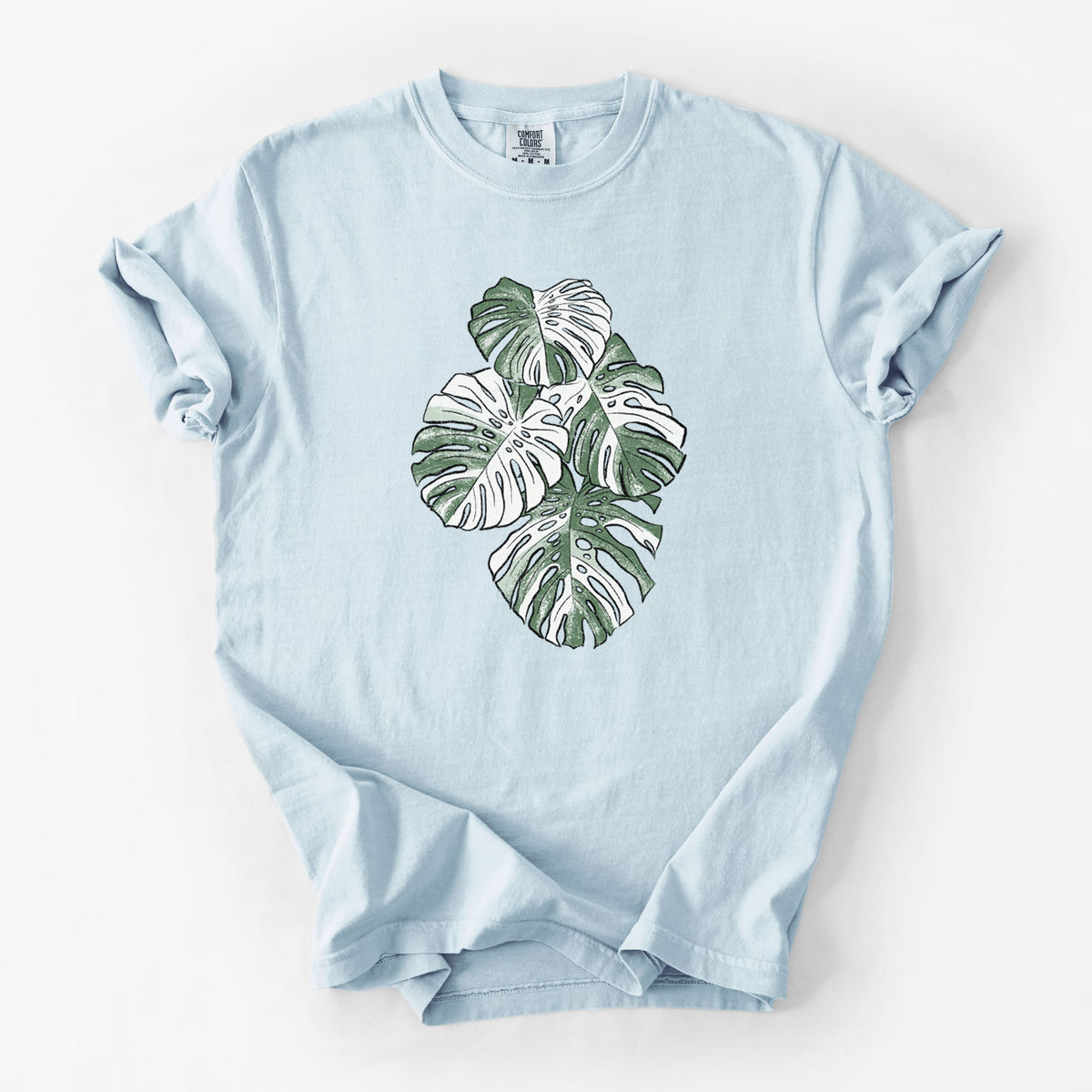 Chambray blue Comfort Colors t-shirt laid flat, printed with a full-color illustration of a variegated Monstera leaf in green and white.