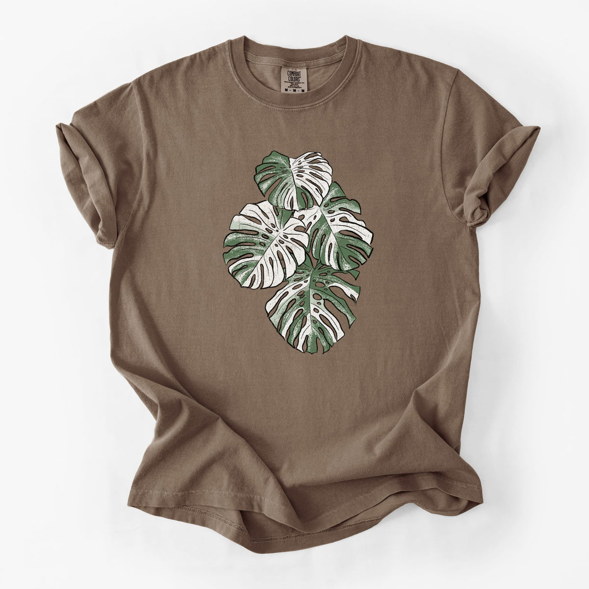 Espresso brown Comfort Colors t-shirt laid flat, printed with a full-color illustration of a variegated Monstera leaf in green and white.
