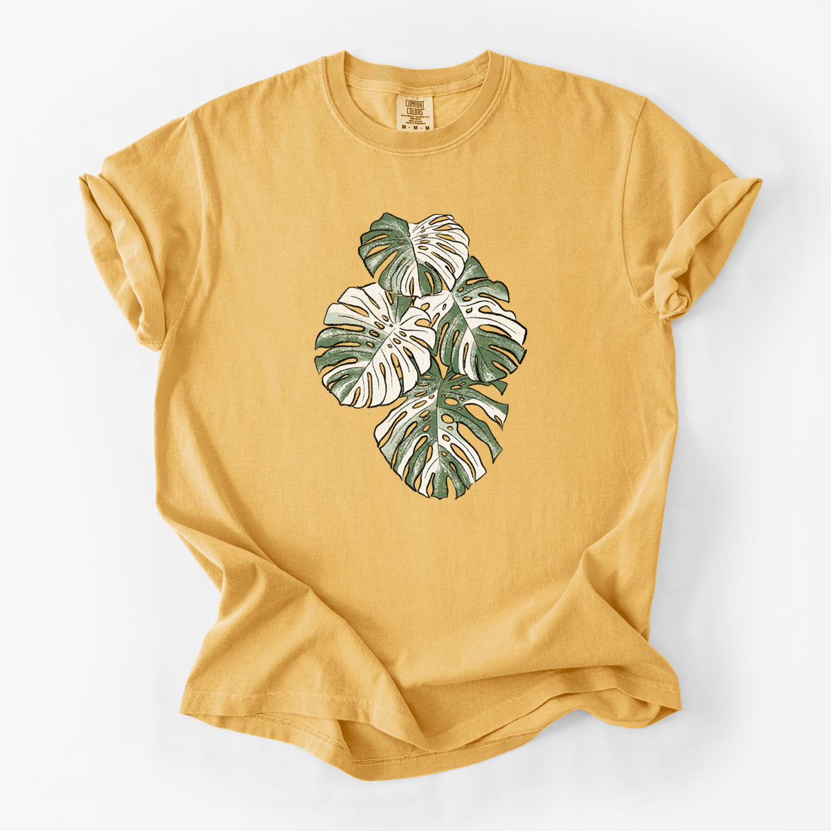 Golden wheat yellow Comfort Colors t-shirt laid flat, printed with a full-color illustration of a variegated Monstera leaf in green and white.