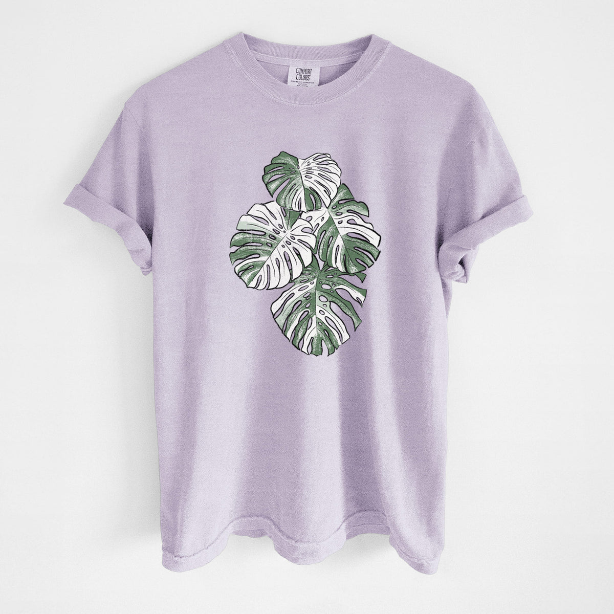 Orchid purple Comfort Colors t-shirt laid flat, printed with a full-color illustration of a variegated Monstera leaf in green and white.