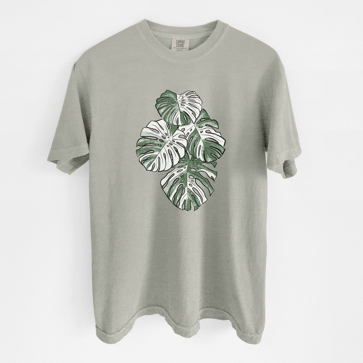Sandstone beige Comfort Colors t-shirt laid flat, printed with a full-color illustration of a variegated Monstera leaf in green and white.