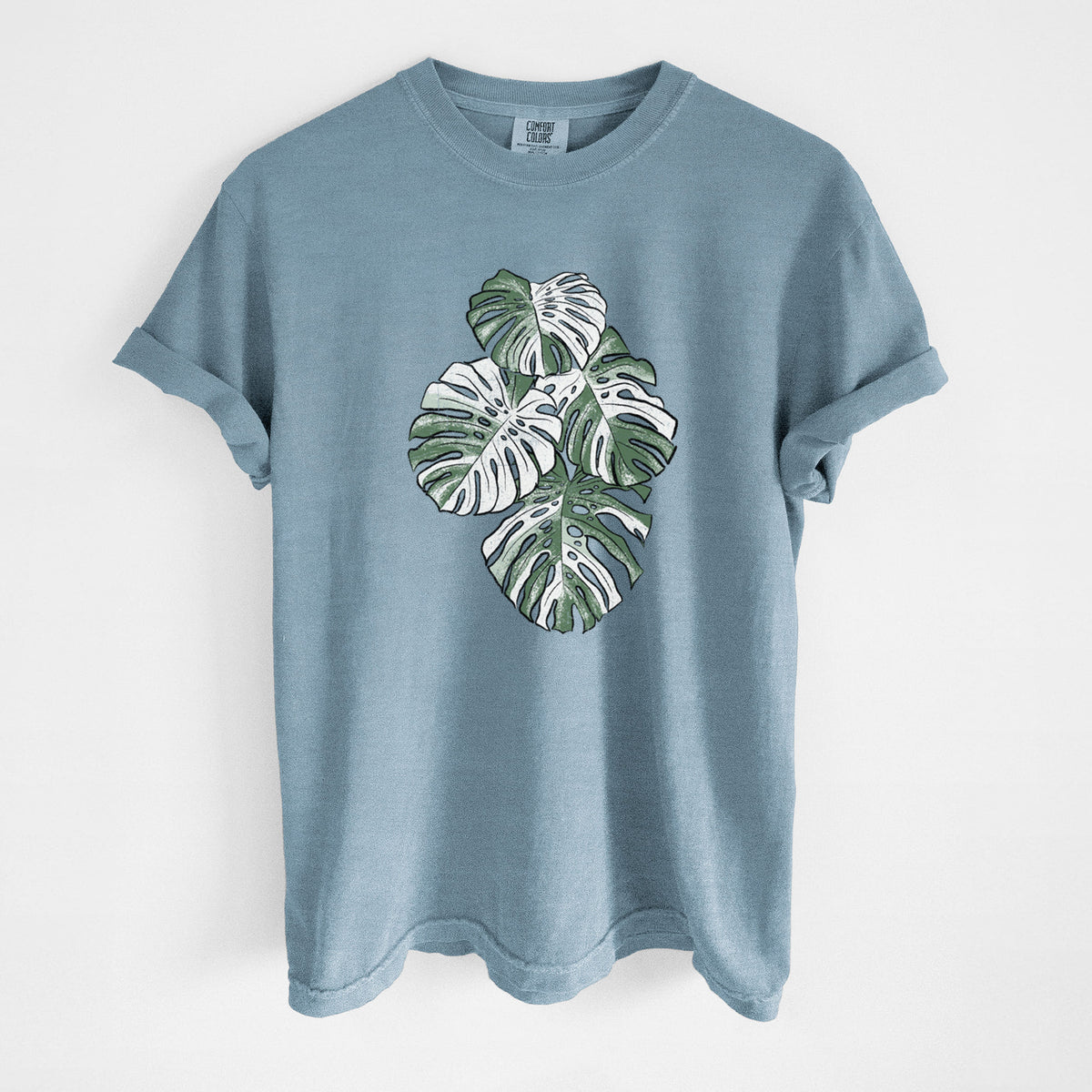 Steel blue Comfort Colors t-shirt laid flat, printed with a full-color illustration of a variegated Monstera leaf in green and white.