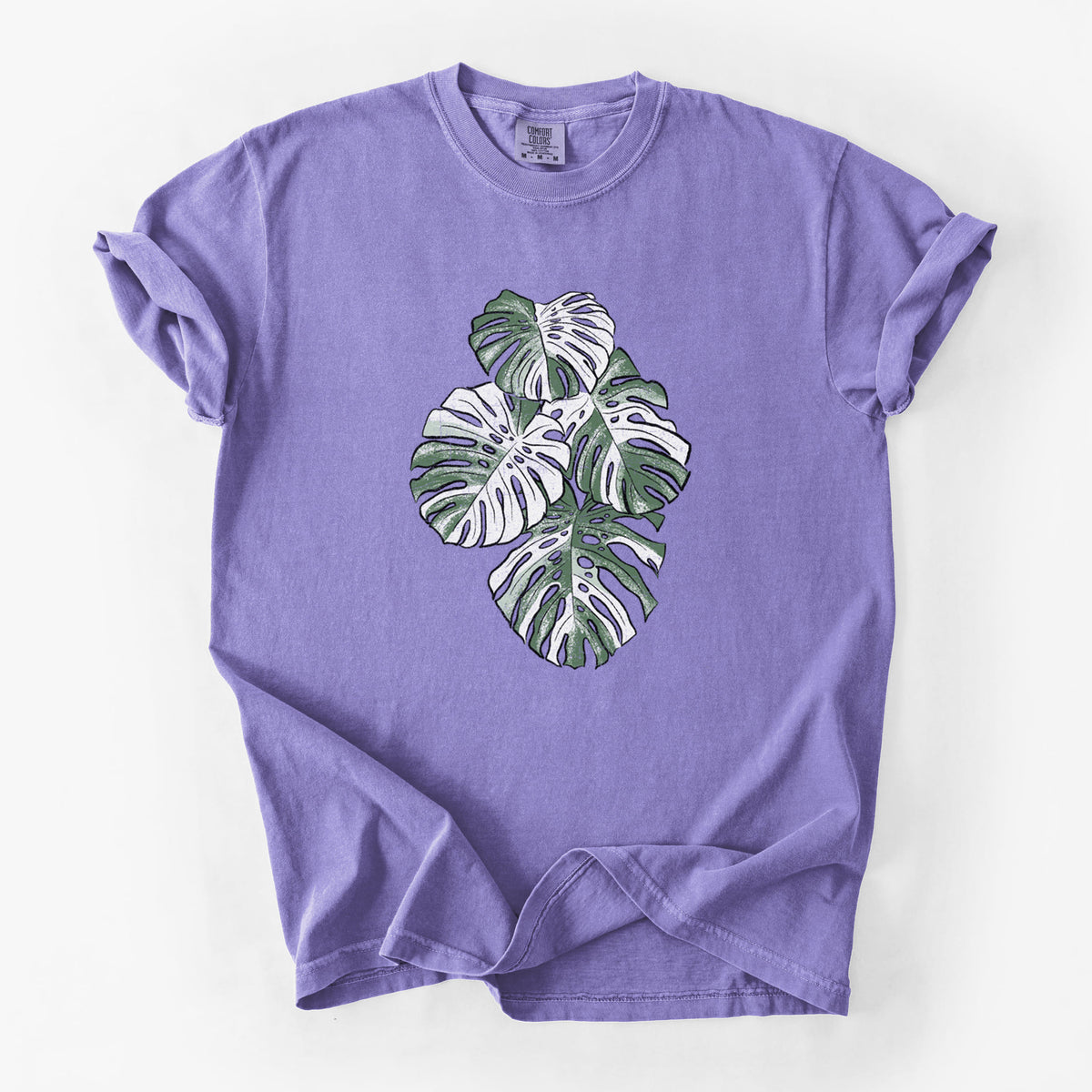 Violet purple Comfort Colors t-shirt laid flat, printed with a full-color illustration of a variegated Monstera leaf in green and white.