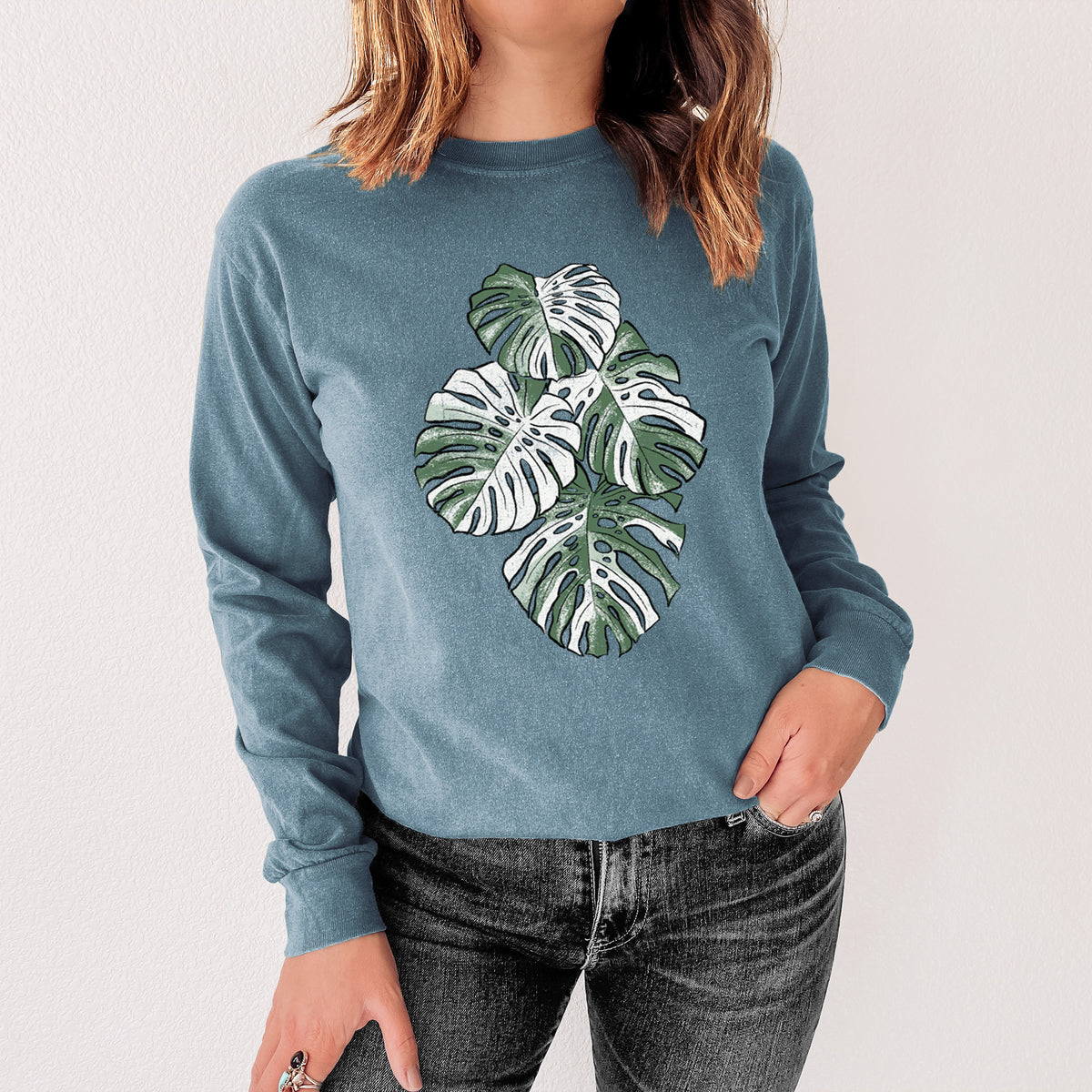 Steel blue Comfort Colors long sleeve shirt printed with a full-color illustration of a variegated Monstera leaf in green and white, shown in a lifestyle photo.