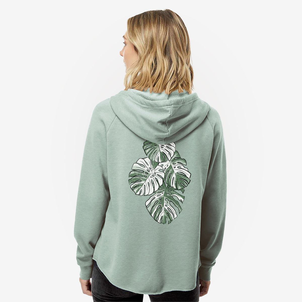 Woman wearing a sage green women's Cali Wave zip-up sweatshirt printed with a full-color illustration of a variegated Monstera leaf in green and white, seen from behind.