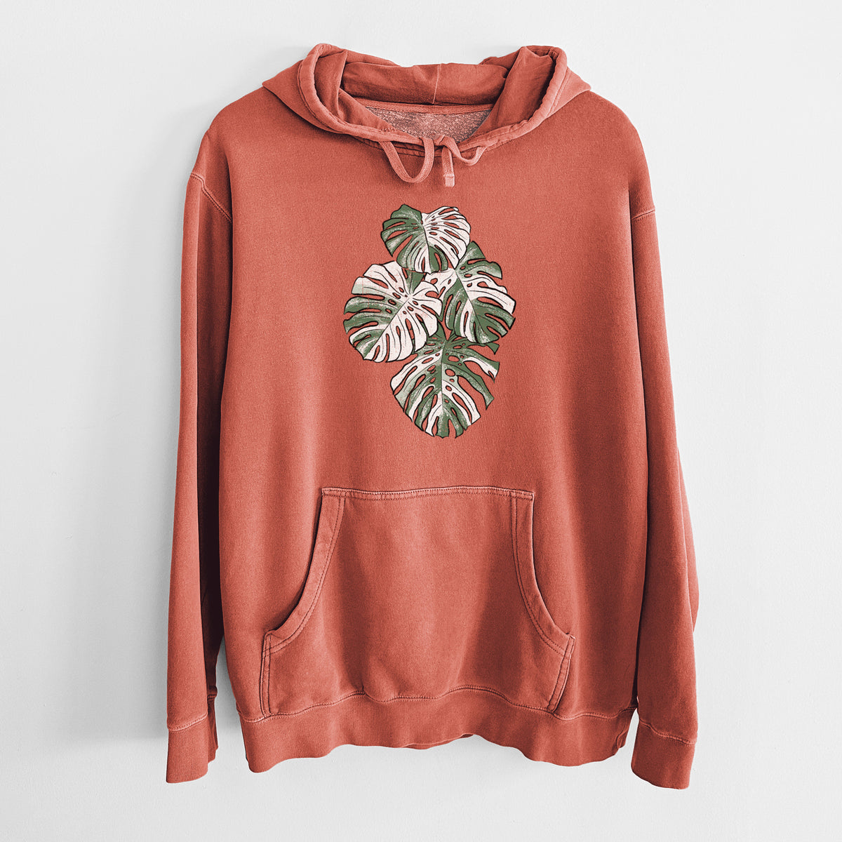 Amber orange pigment dyed hoodie laid flat, printed with a full-color illustration of a variegated Monstera leaf in green and white.