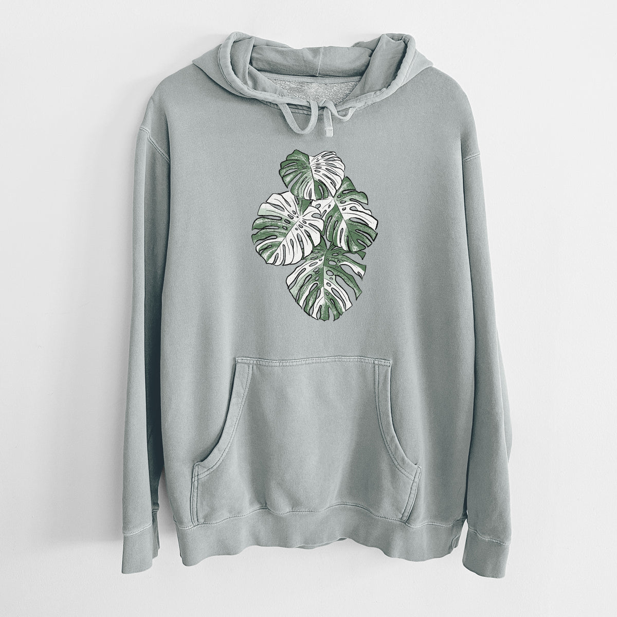Sage green pigment dyed hoodie laid flat, printed with a full-color illustration of a variegated Monstera leaf in green and white.
