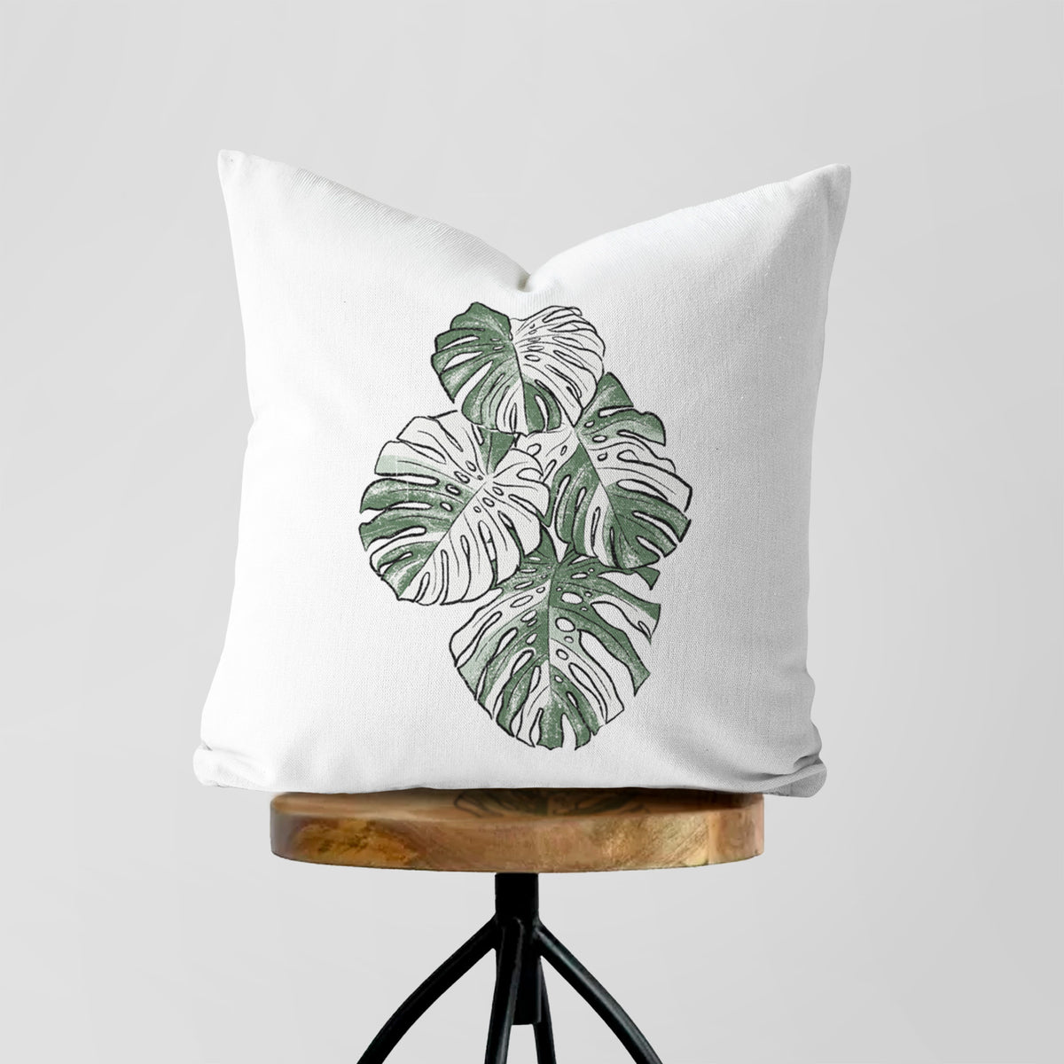 White pillow cover printed with a full-color illustration of a variegated Monstera leaf in green and white, on a wooden stool.