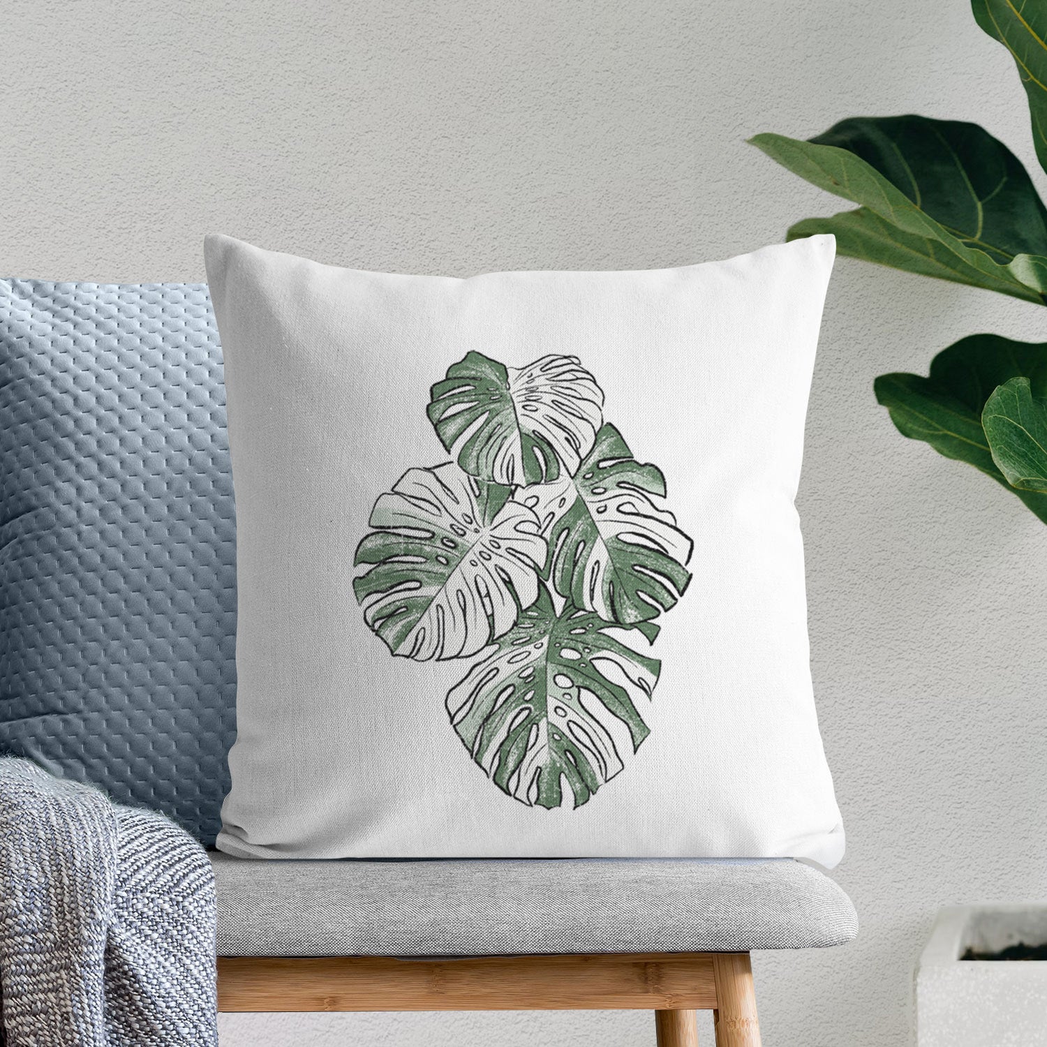 White pillow cover printed with a full-color illustration of a variegated Monstera leaf in green and white, on a bench beside plants.