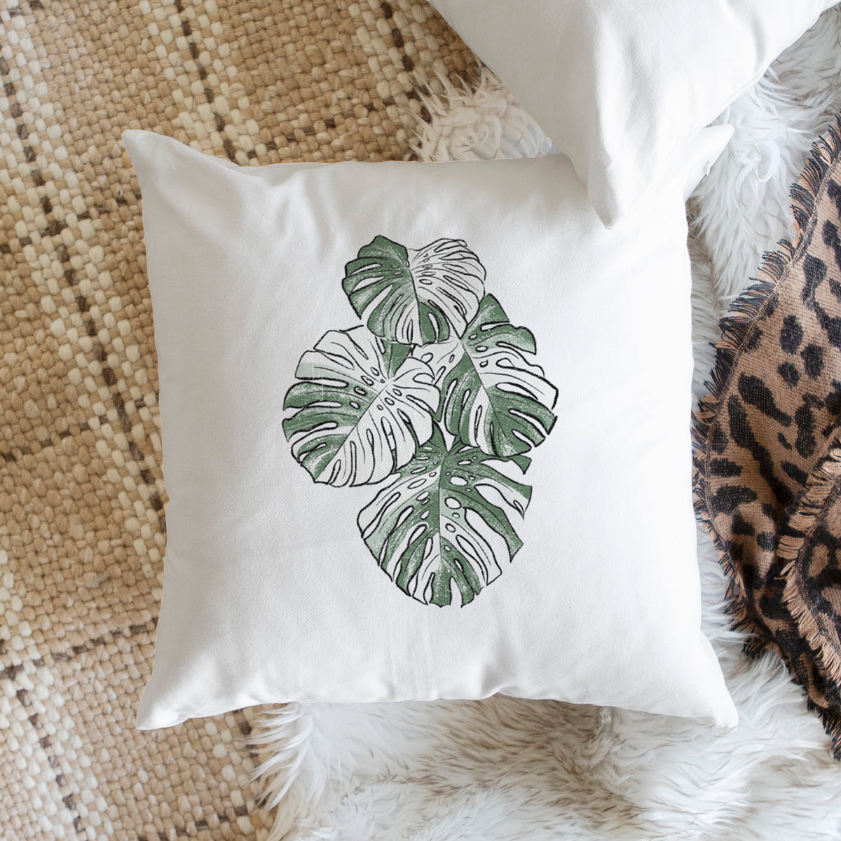 White pillow cover printed with a full-color illustration of a variegated Monstera leaf in green and white, on a woven chair.