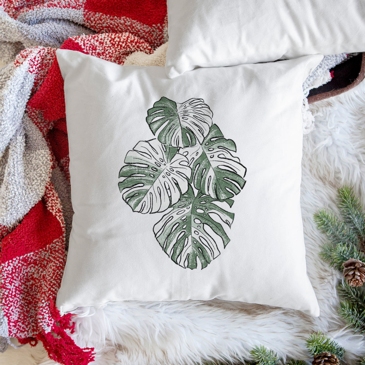 White pillow cover printed with a full-color illustration of a variegated Monstera leaf in green and white, on a woven chair.