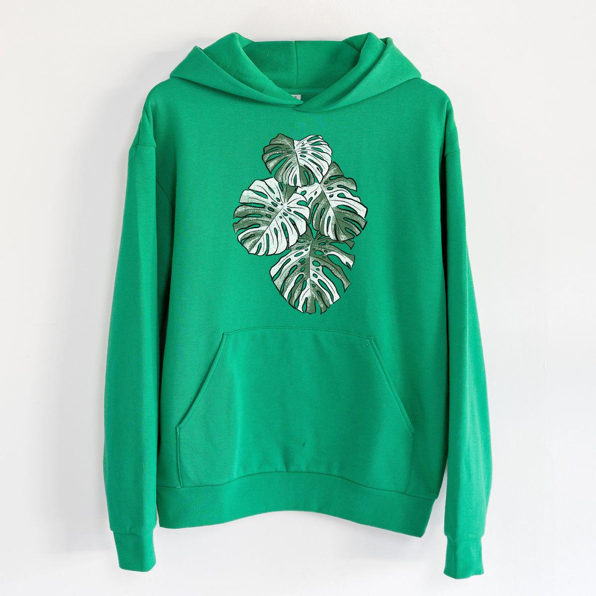 Variegated Monstera - Statement Midweight Hoodie in Kelly Green, hanging on a white wall, with a front kangaroo pocket and no drawcords.