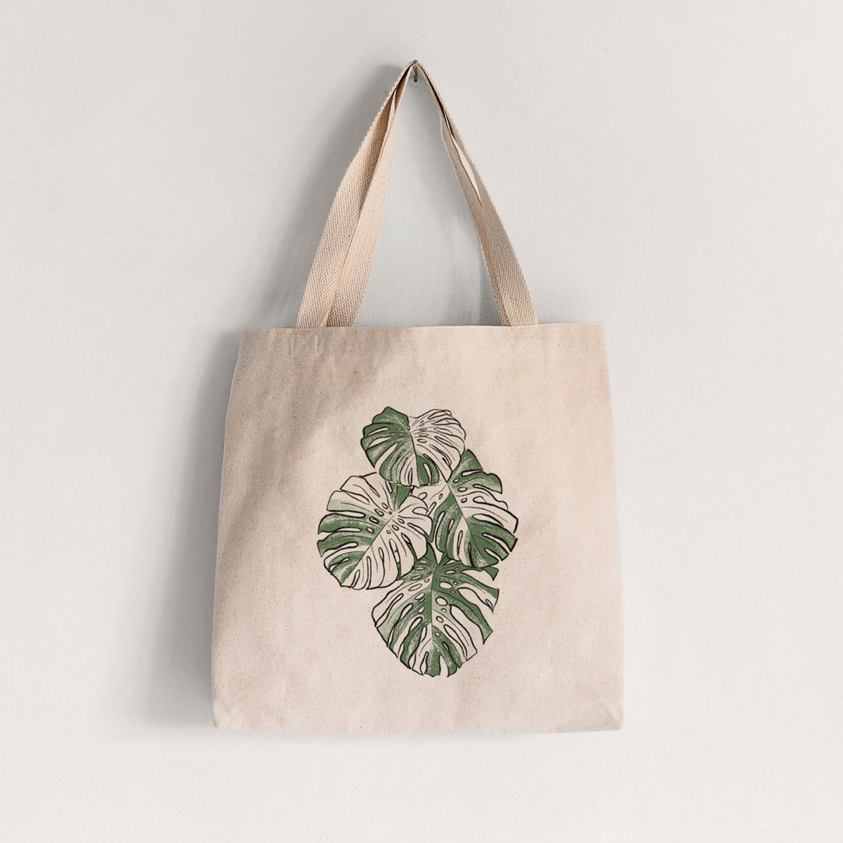 Natural canvas tote bag printed with a full-color illustration of a variegated Monstera leaf in green and white.
