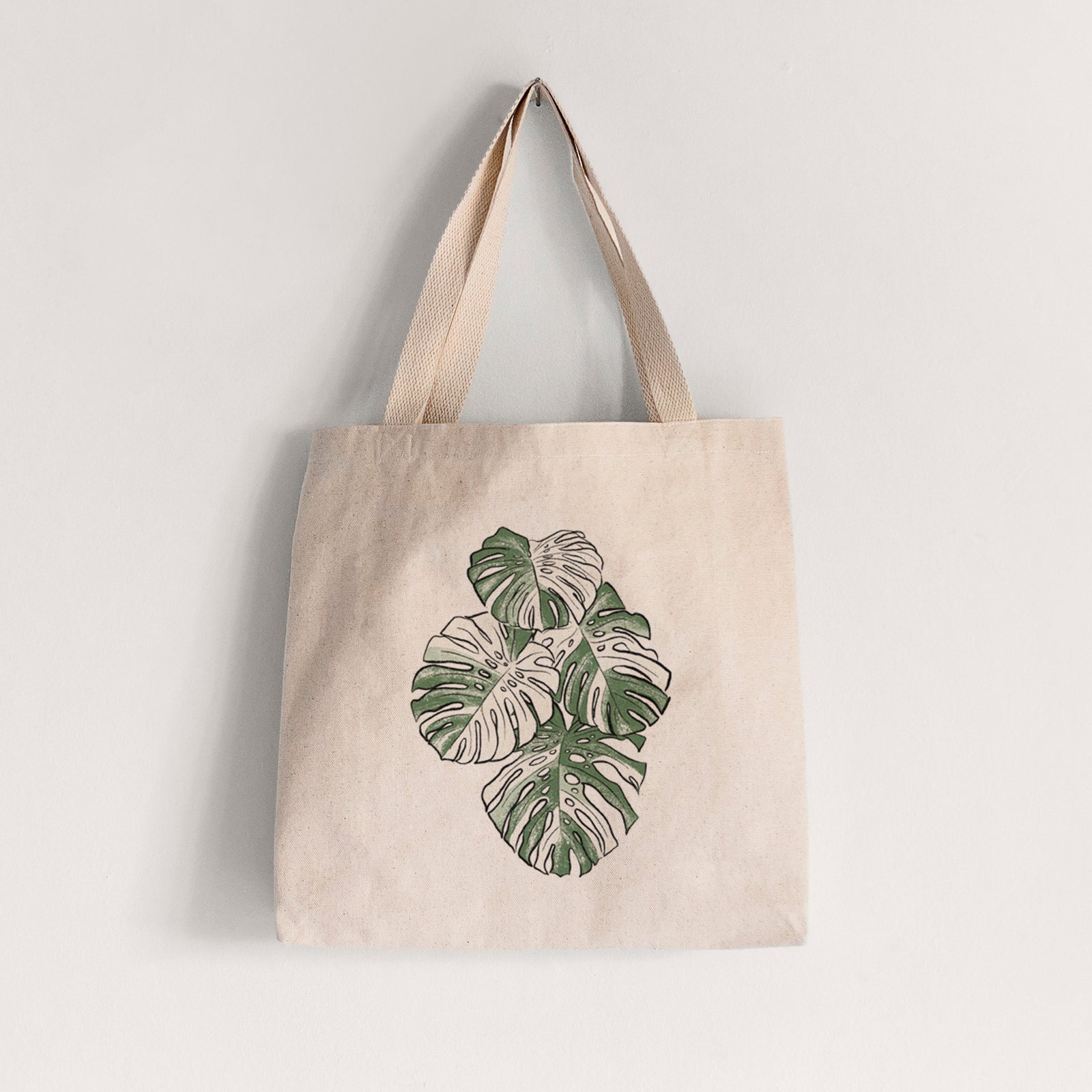 Natural canvas tote bag printed with a full-color illustration of a variegated Monstera leaf in green and white.