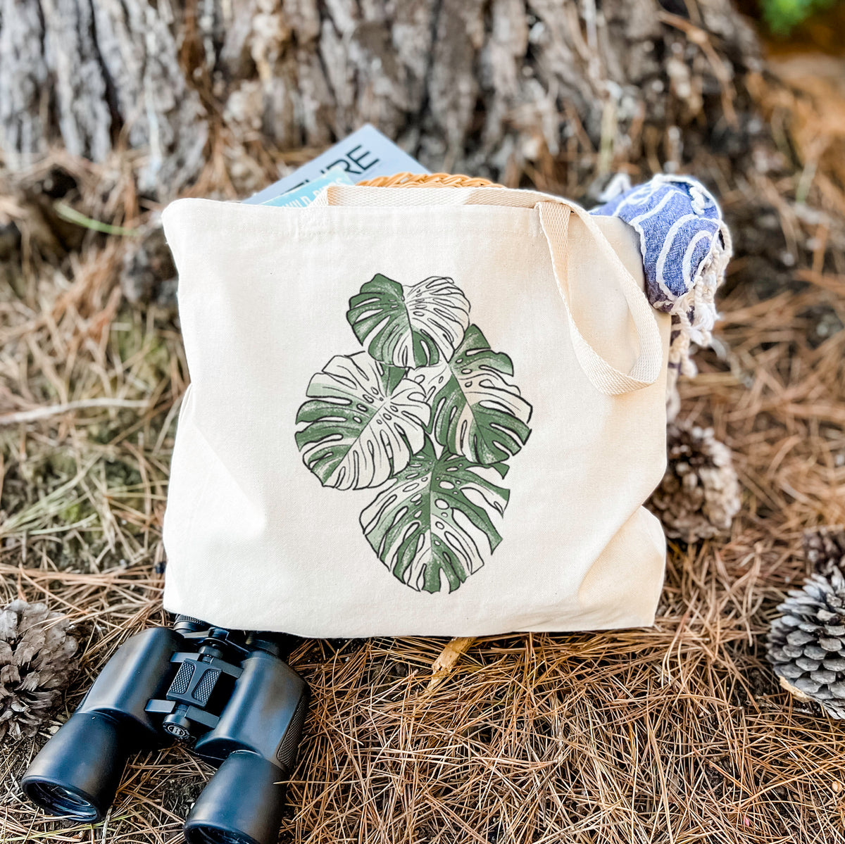 Canvas tote bag printed with a full-color illustration of a variegated Monstera leaf in green and white, resting on dry grass beside a pair of binoculars.