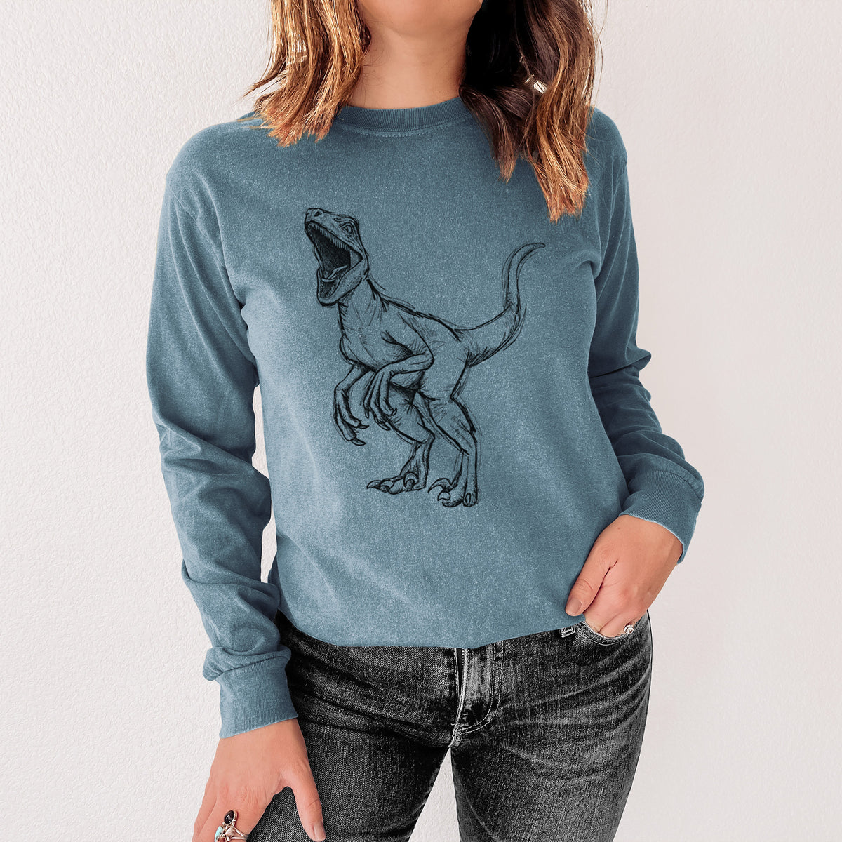 Steel blue Comfort Colors long sleeve shirt printed with a black ink illustration of a Velociraptor roaring, shown in a lifestyle photo.