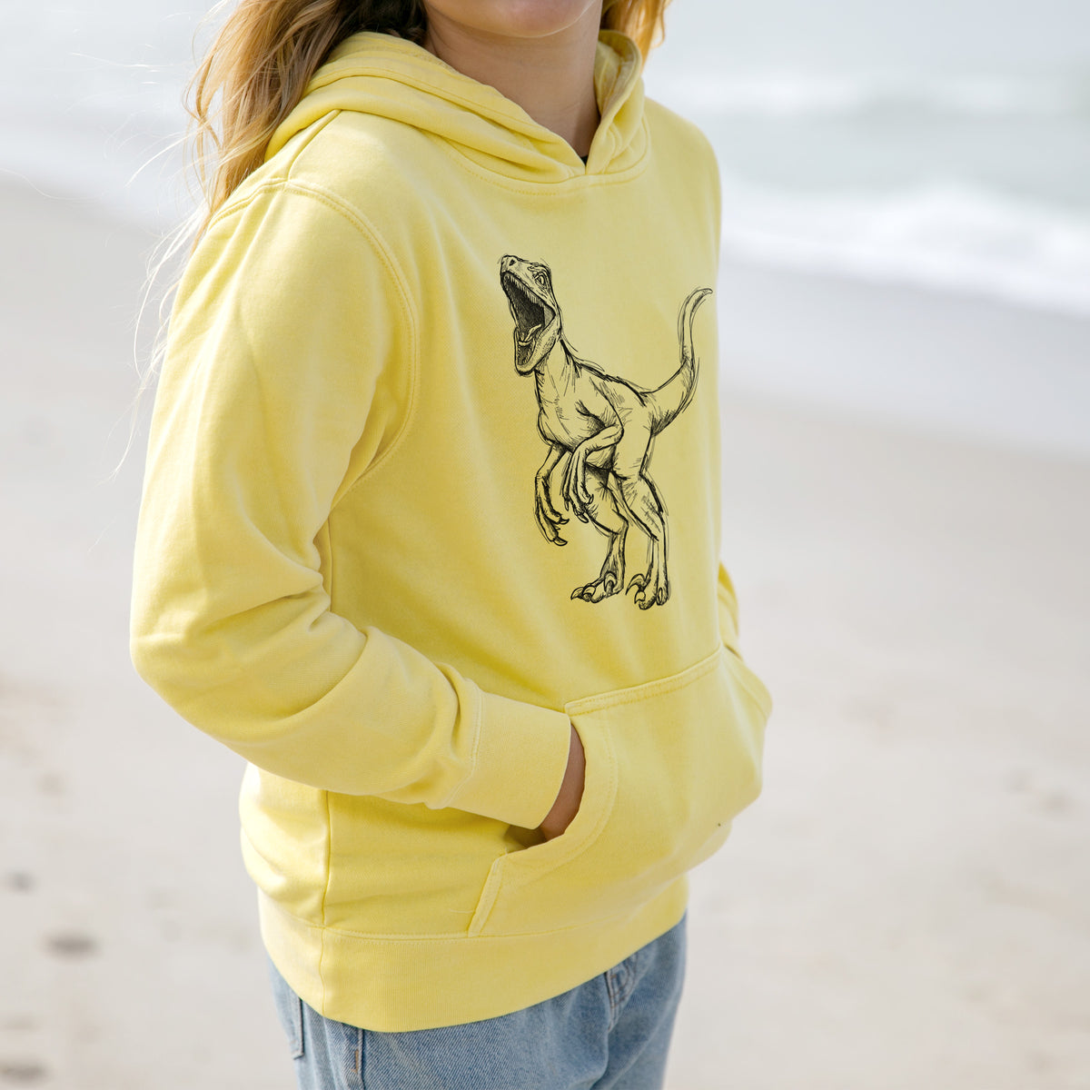 Girl wearing a youth pigment dyed hoodie printed with a black ink illustration of a Velociraptor roaring.