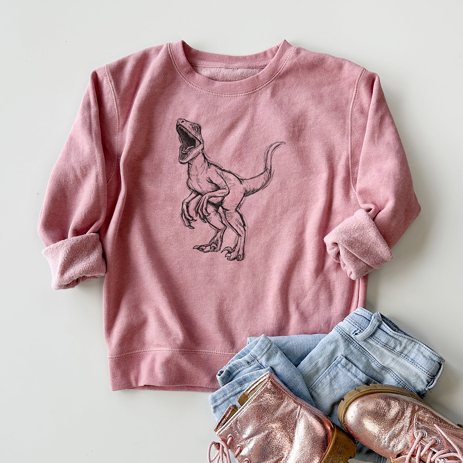 Boy wearing a youth crewneck sweatshirt printed with a black ink illustration of a Velociraptor roaring, outdoors.