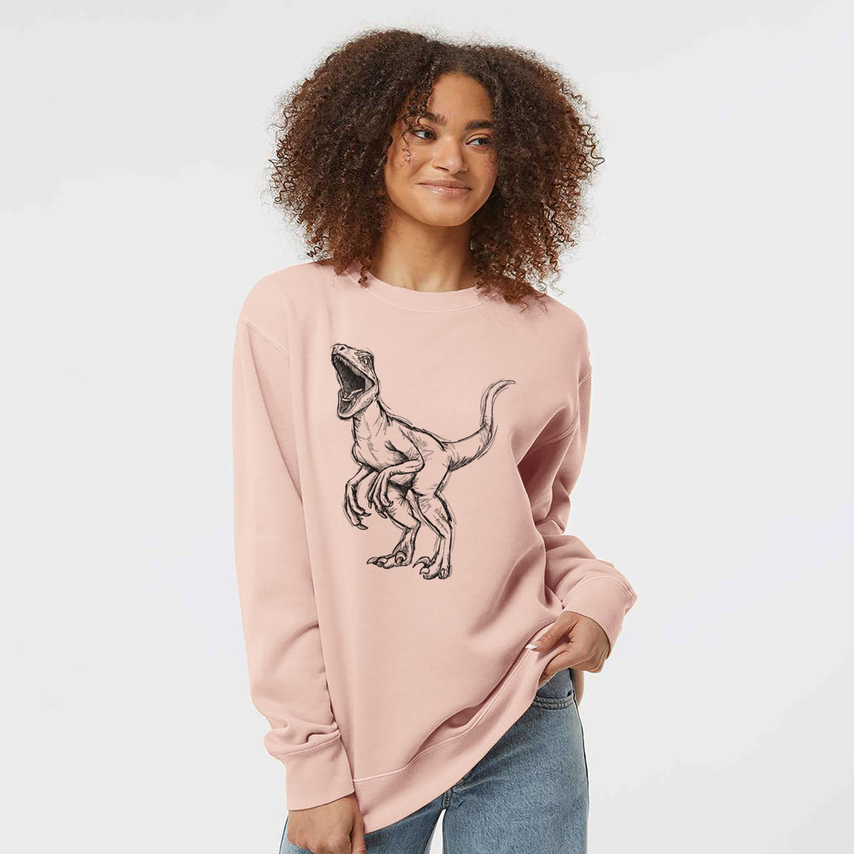 Model wearing a dusty pink pigment dyed crewneck sweatshirt printed with a black ink illustration of a Velociraptor roaring.