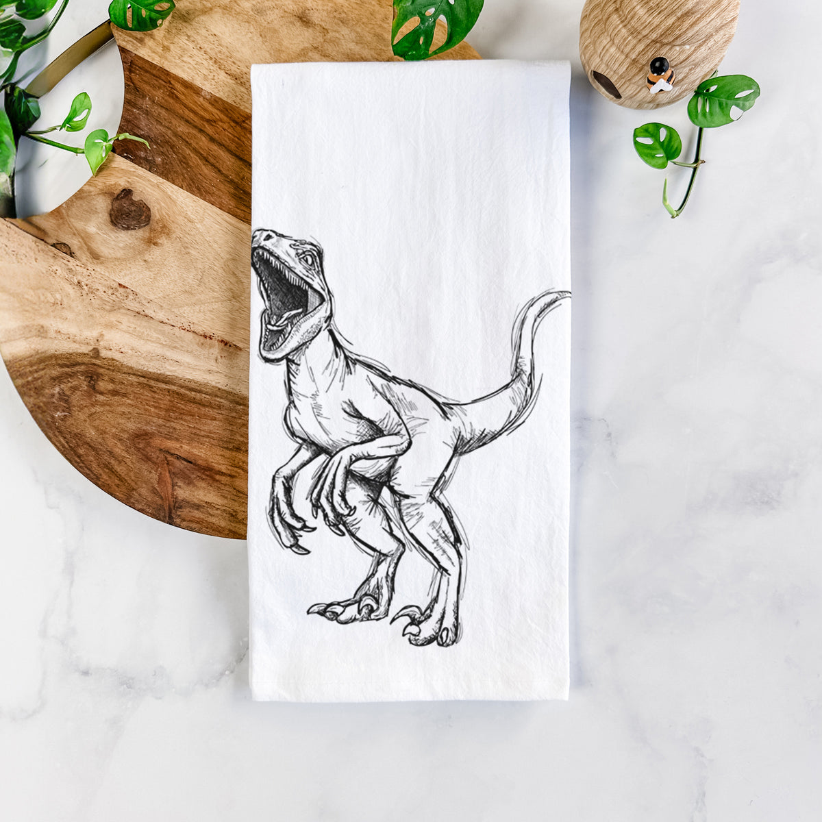 White cotton tea towel printed with a black ink illustration of a Velociraptor roaring, on a marble counter beside a round wooden cutting board and greenery.