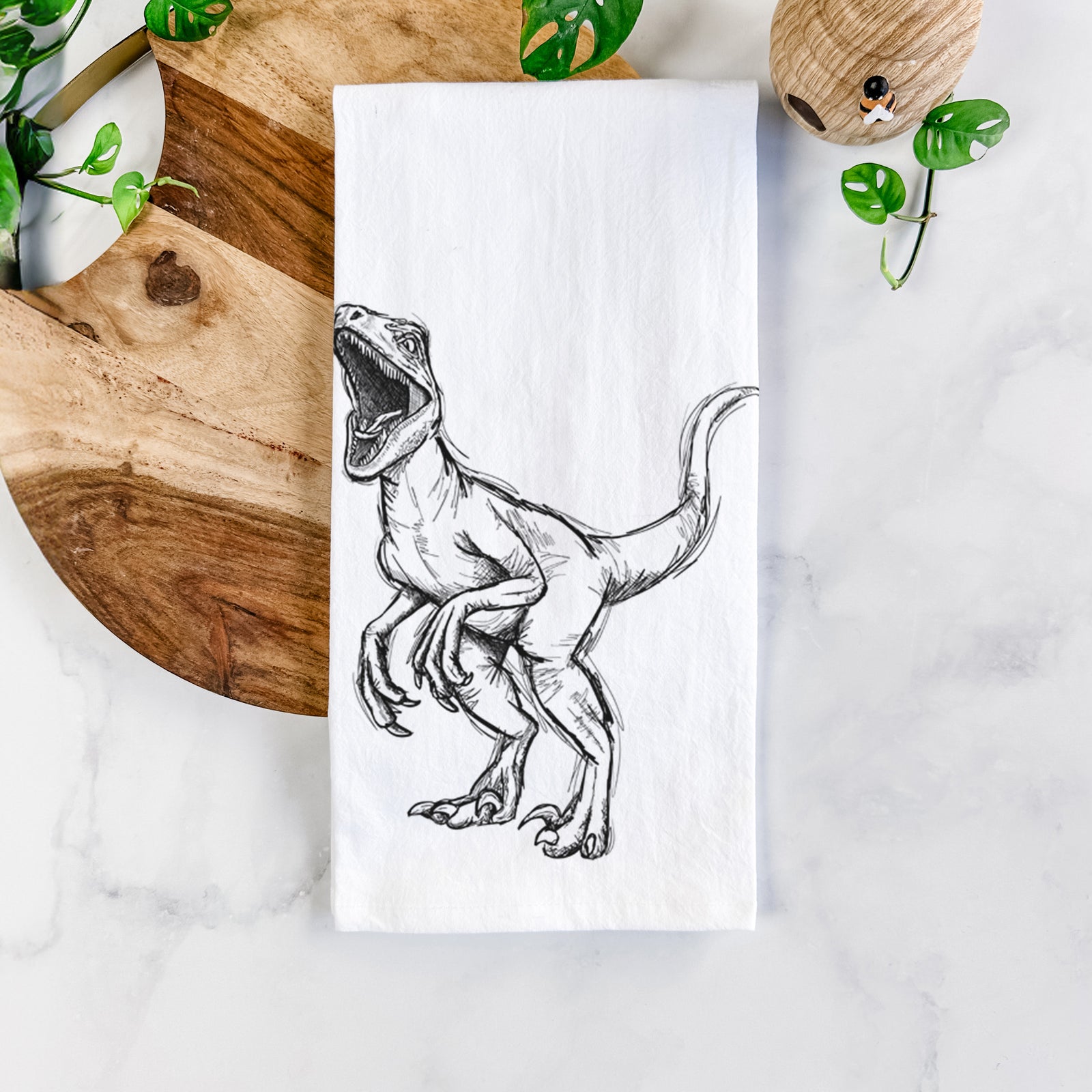White cotton tea towel printed with a black ink illustration of a Velociraptor roaring, on a marble counter beside a round wooden cutting board and greenery.