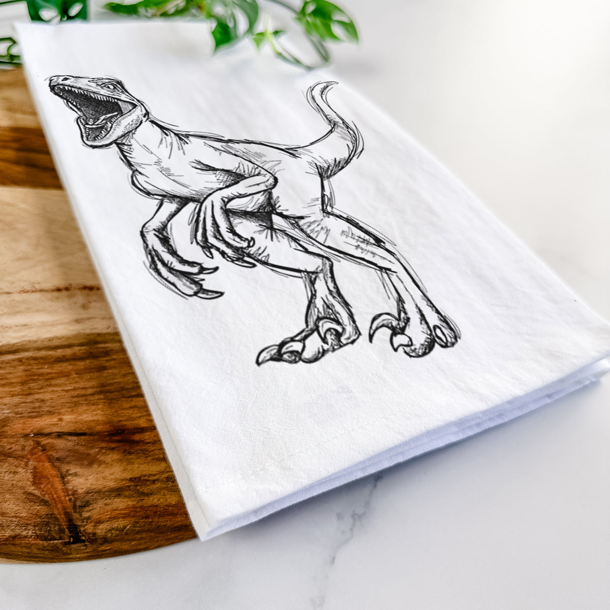 Close-up of a folded white tea towel on a wooden board, showing a black ink illustration of a Velociraptor roaring.