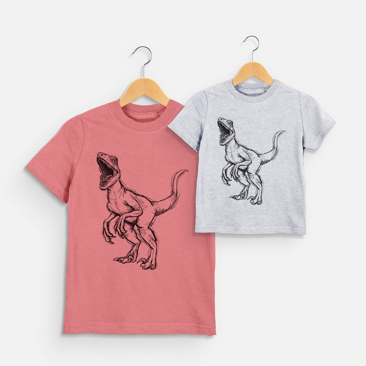 Two youth t-shirts in different colors on hangers, printed with a black ink illustration of a Velociraptor roaring.