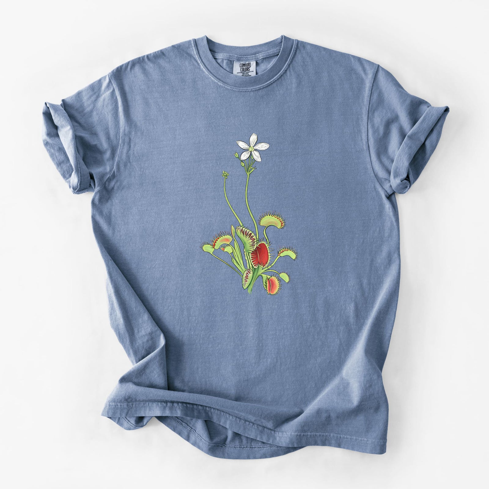 Blue jean Comfort Colors t-shirt laid flat, printed with a full-color illustration of a Venus flytrap plant with red traps and a small white flower.