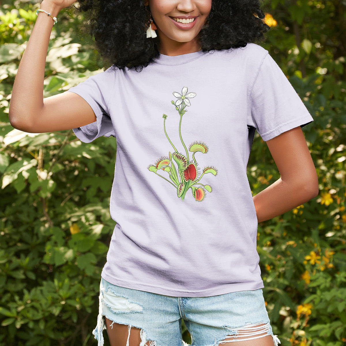 Woman wearing a Comfort Colors t-shirt printed with a full-color illustration of a Venus flytrap plant with red traps and a small white flower, outdoors.