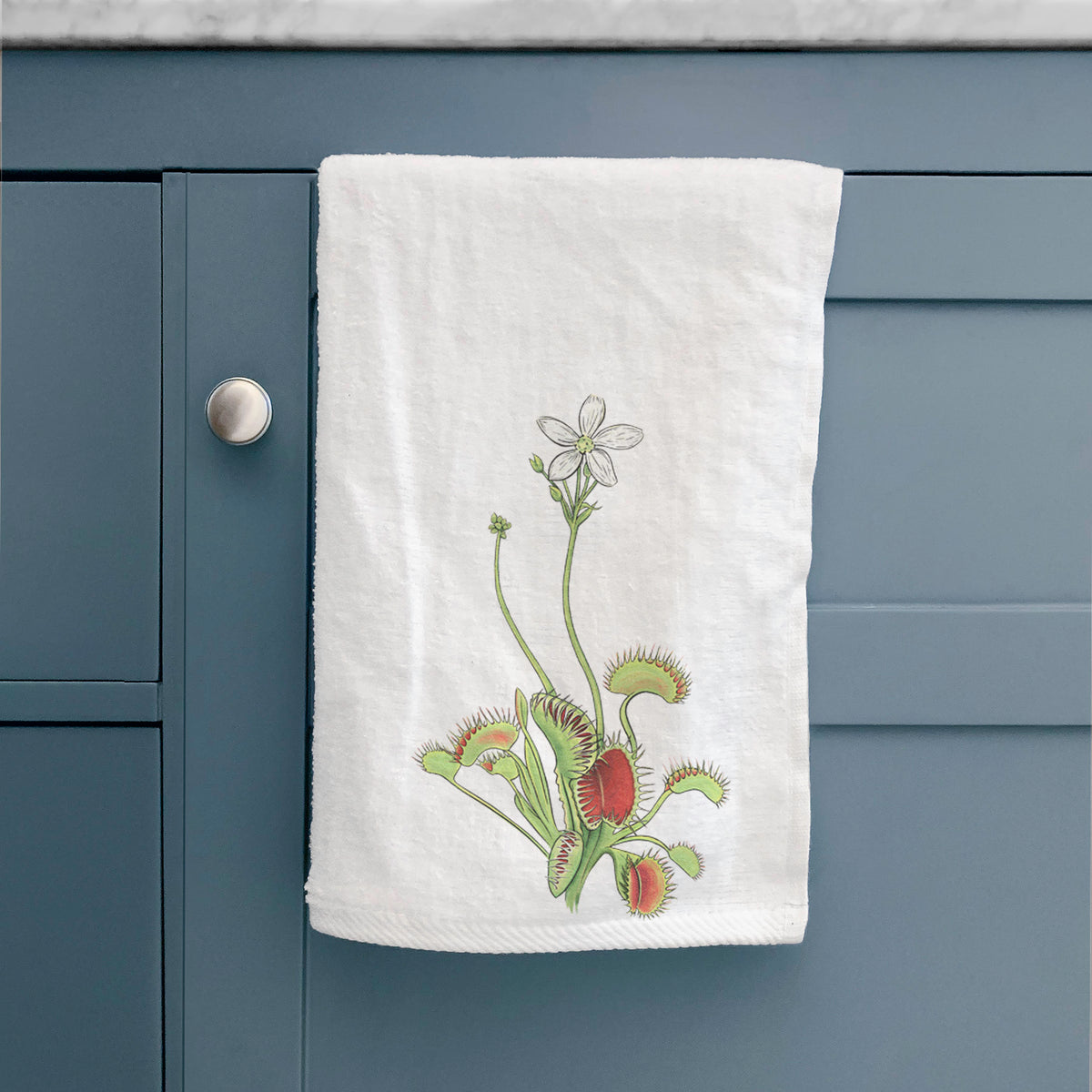 White hand towel printed with a full-color illustration of a Venus flytrap plant with red traps and a small white flower, hanging on a cabinet door.