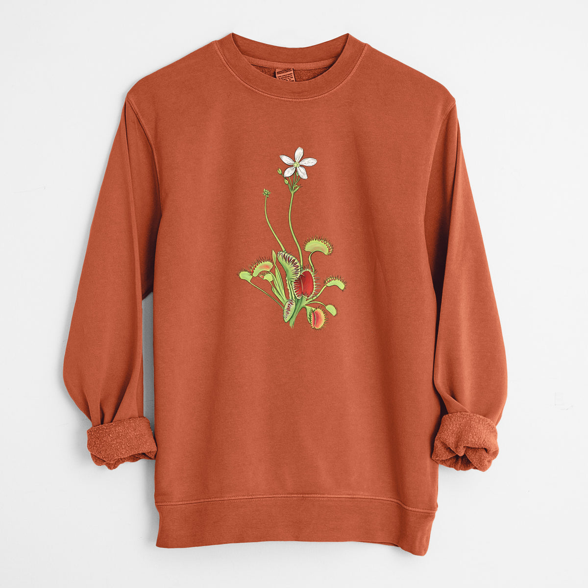 Amber orange pigment dyed crewneck sweatshirt laid flat with the sleeves rolled, printed with a full-color illustration of a Venus flytrap plant with red traps and a small white flower.