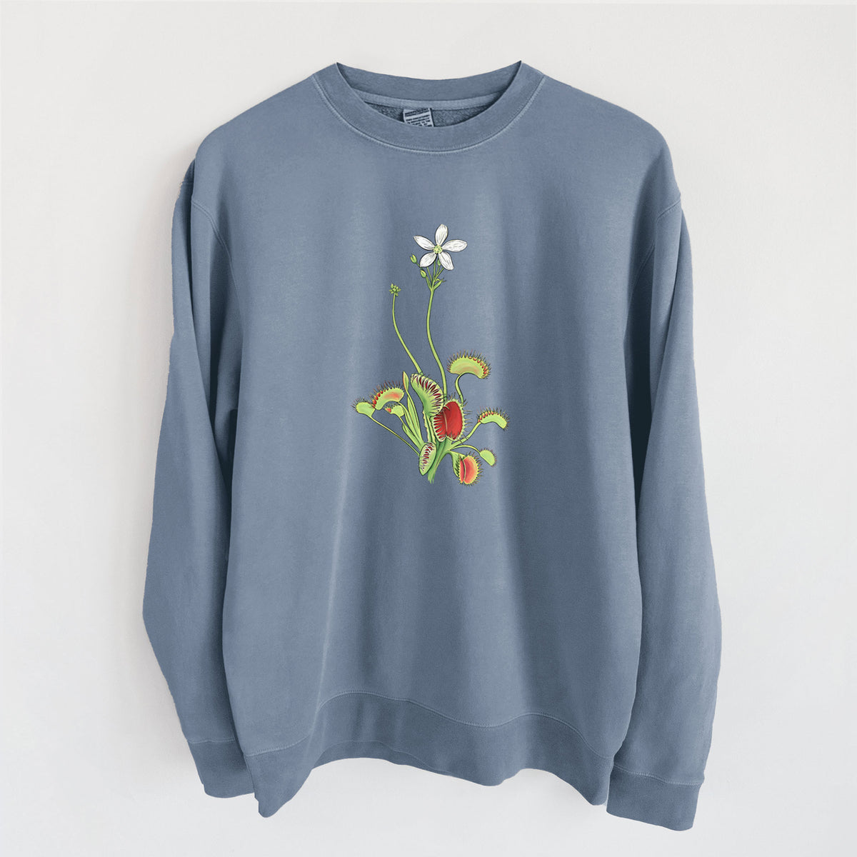 Slate blue pigment dyed crewneck sweatshirt laid flat, printed with a full-color illustration of a Venus flytrap plant with red traps and a small white flower.