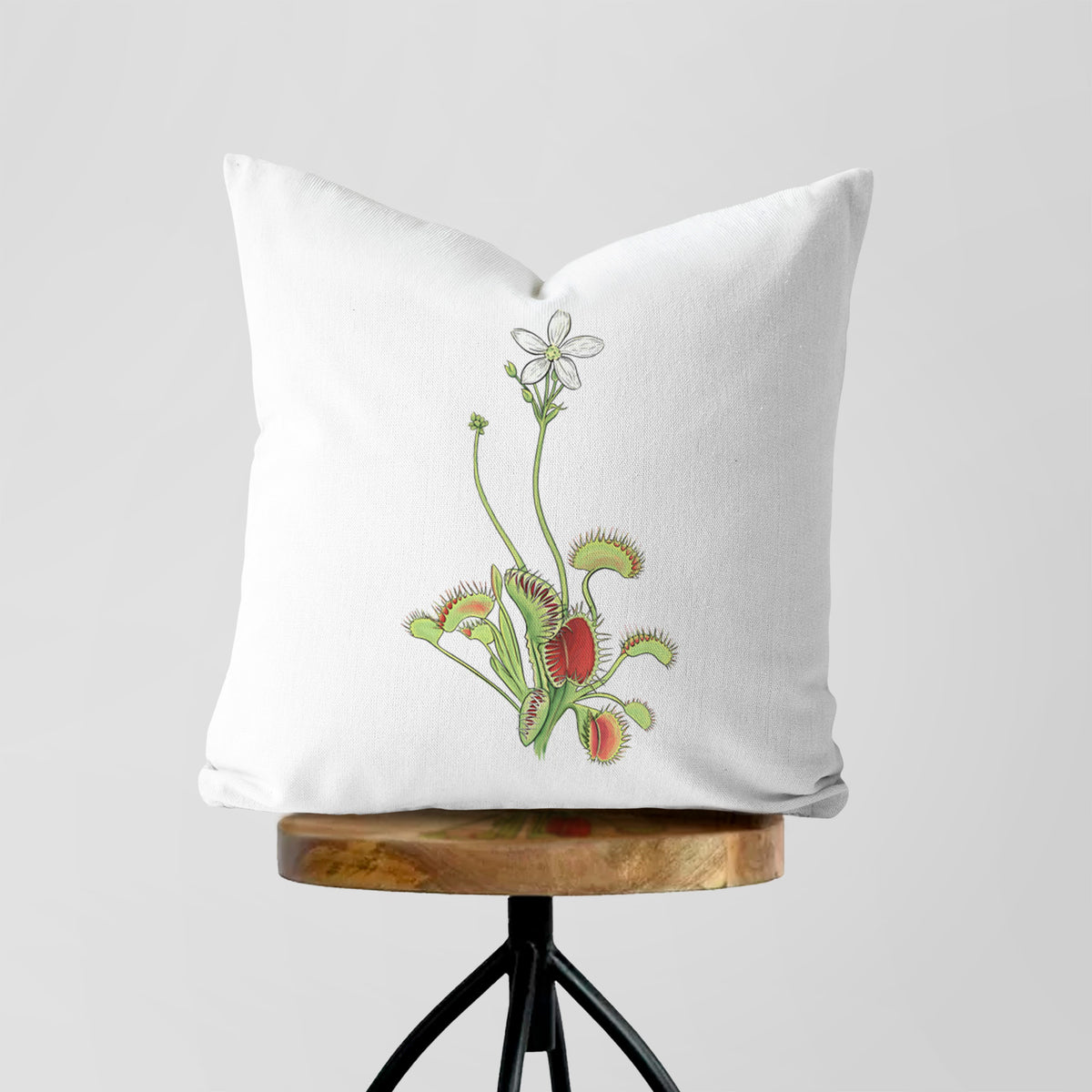 White pillow cover printed with a full-color illustration of a Venus flytrap plant with red traps and a small white flower, on a wooden stool.