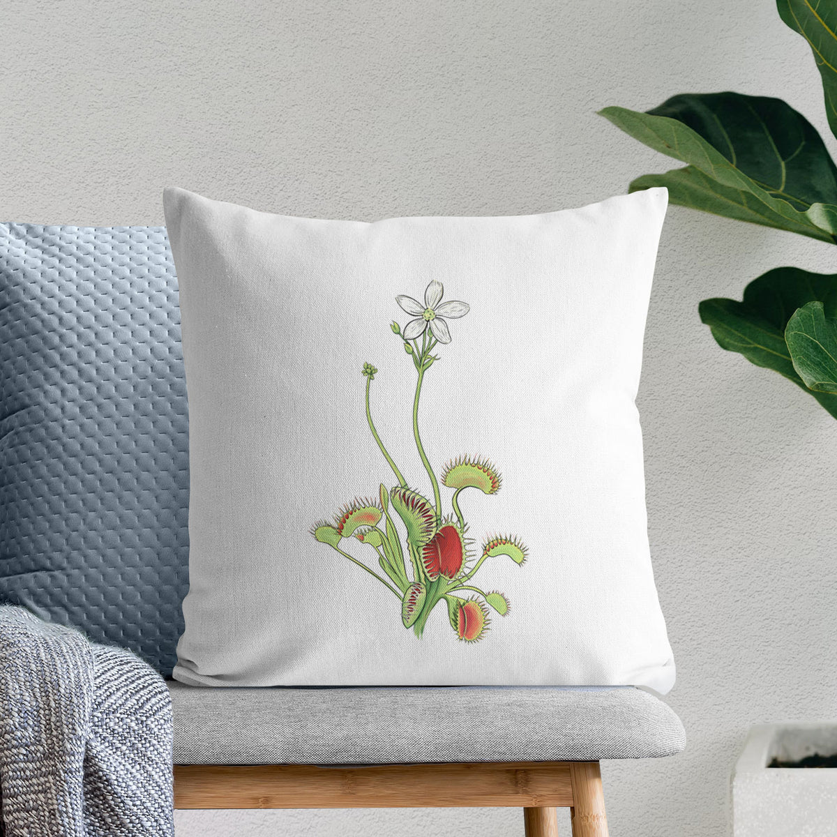 White pillow cover printed with a full-color illustration of a Venus flytrap plant with red traps and a small white flower, on a bench beside plants.